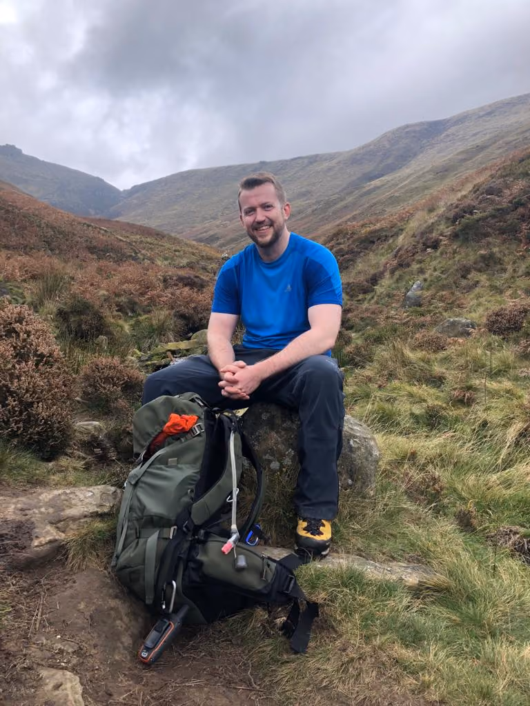 Man in blue shirt and dark pants sitting on a rock in a hilly, grassy landscape with a large green backpack on the ground beside him under an overcast sky.