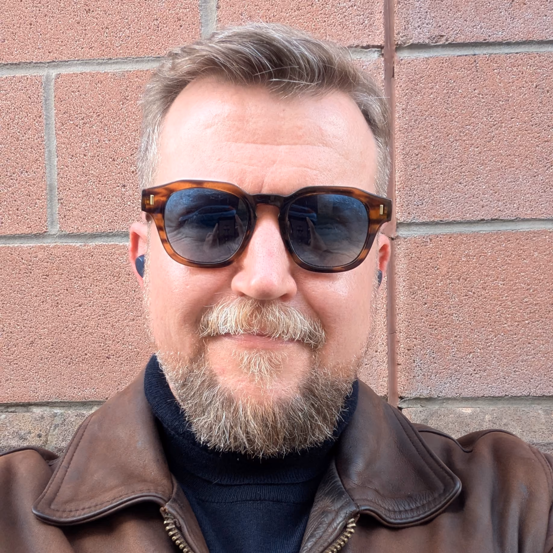 Close-up of a man with light brown hair and beard wearing tortoiseshell sunglasses and a brown leather jacket over a black turtleneck, standing against a pinkish brick wall.
