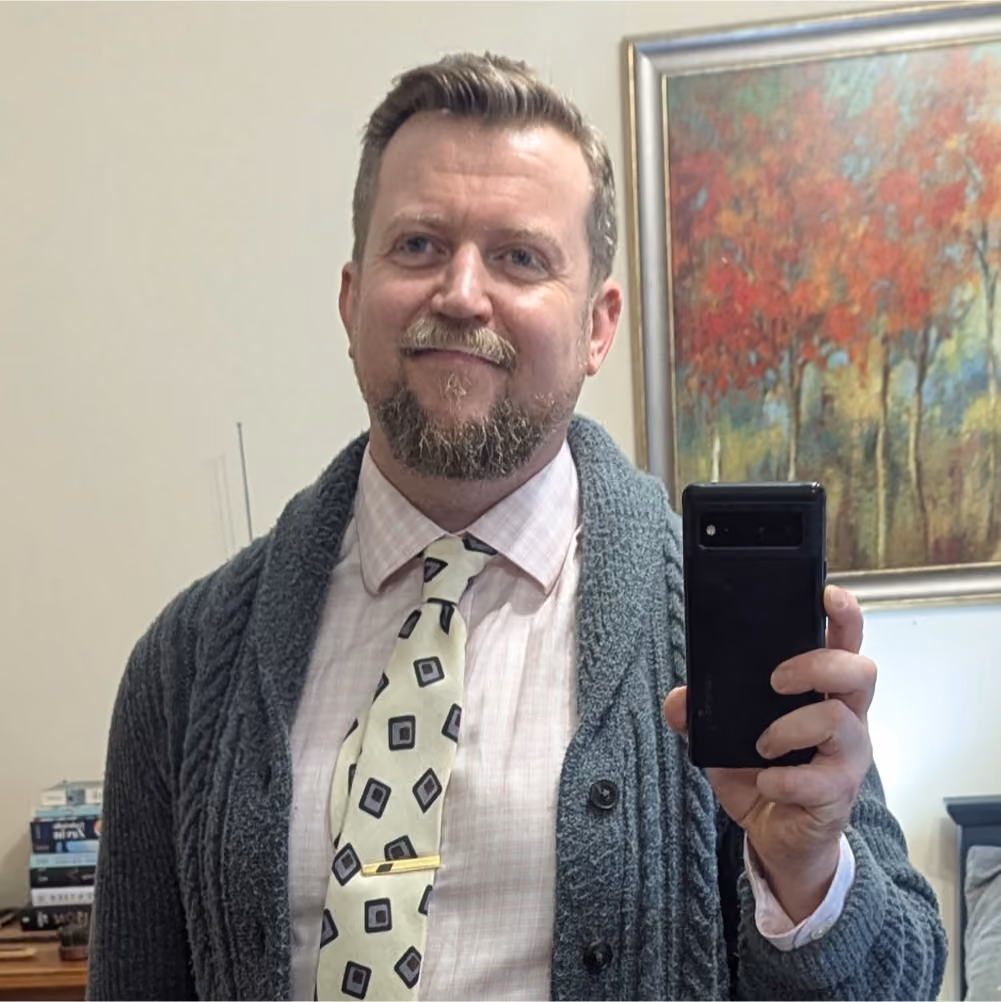 Man with short hair and beard wearing a light pink checkered shirt, cream tie with black squares, and chunky gray cardigan taking a mirror selfie with a black smartphone, with a colorful autumnal tree painting in the background.