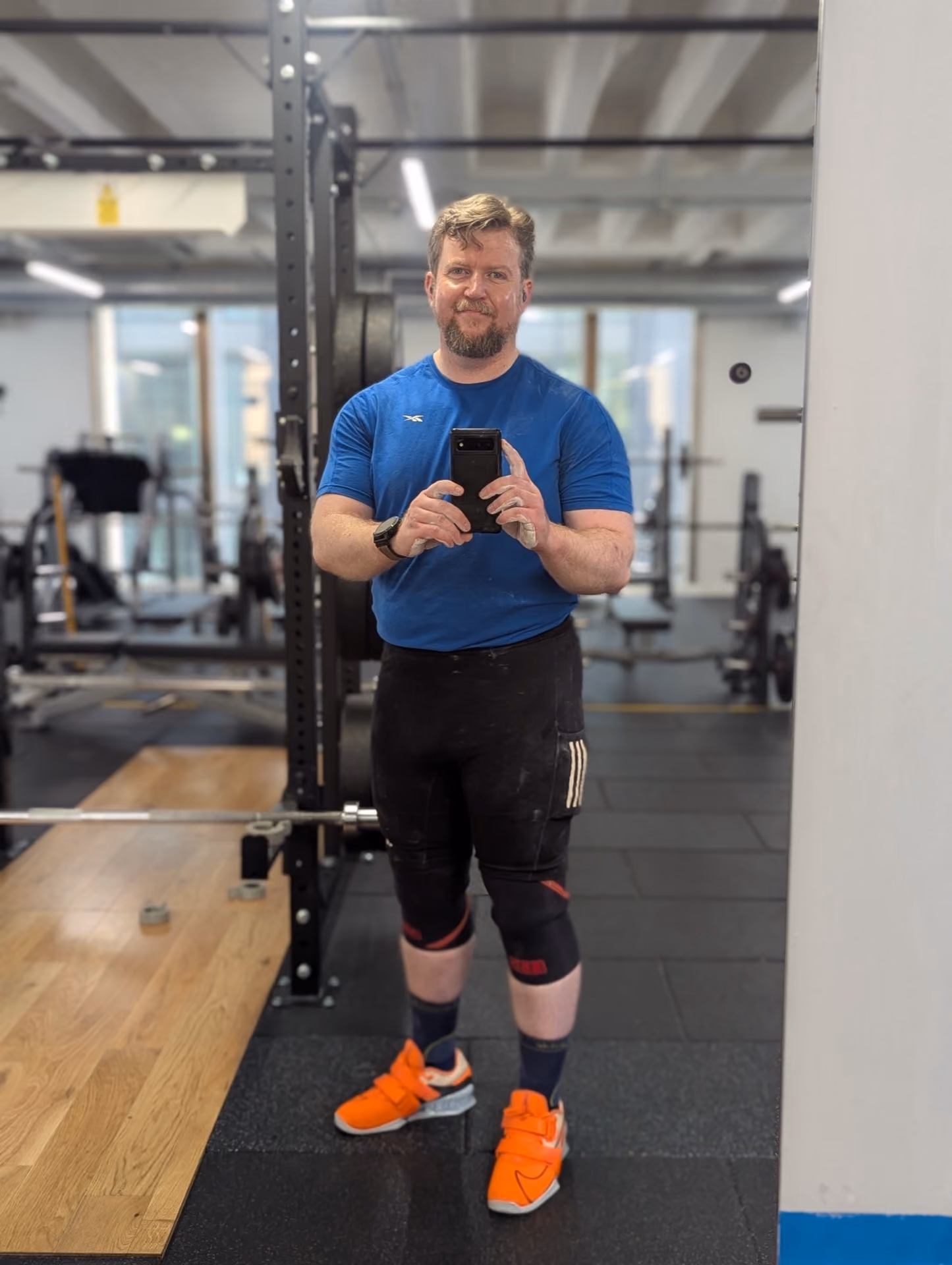 A man with a beard and short hair taking a selfie in a gym mirror, wearing a blue shirt, black leggings, knee sleeves, and bright orange athletic shoes, surrounded by gym equipment.