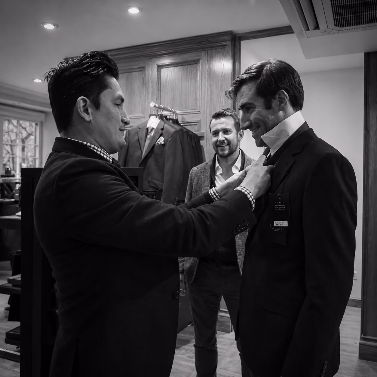 Two men in a suit shop with one man adjusting the tie of another man wearing a suit with a price tag, while a third man stands smiling behind them.